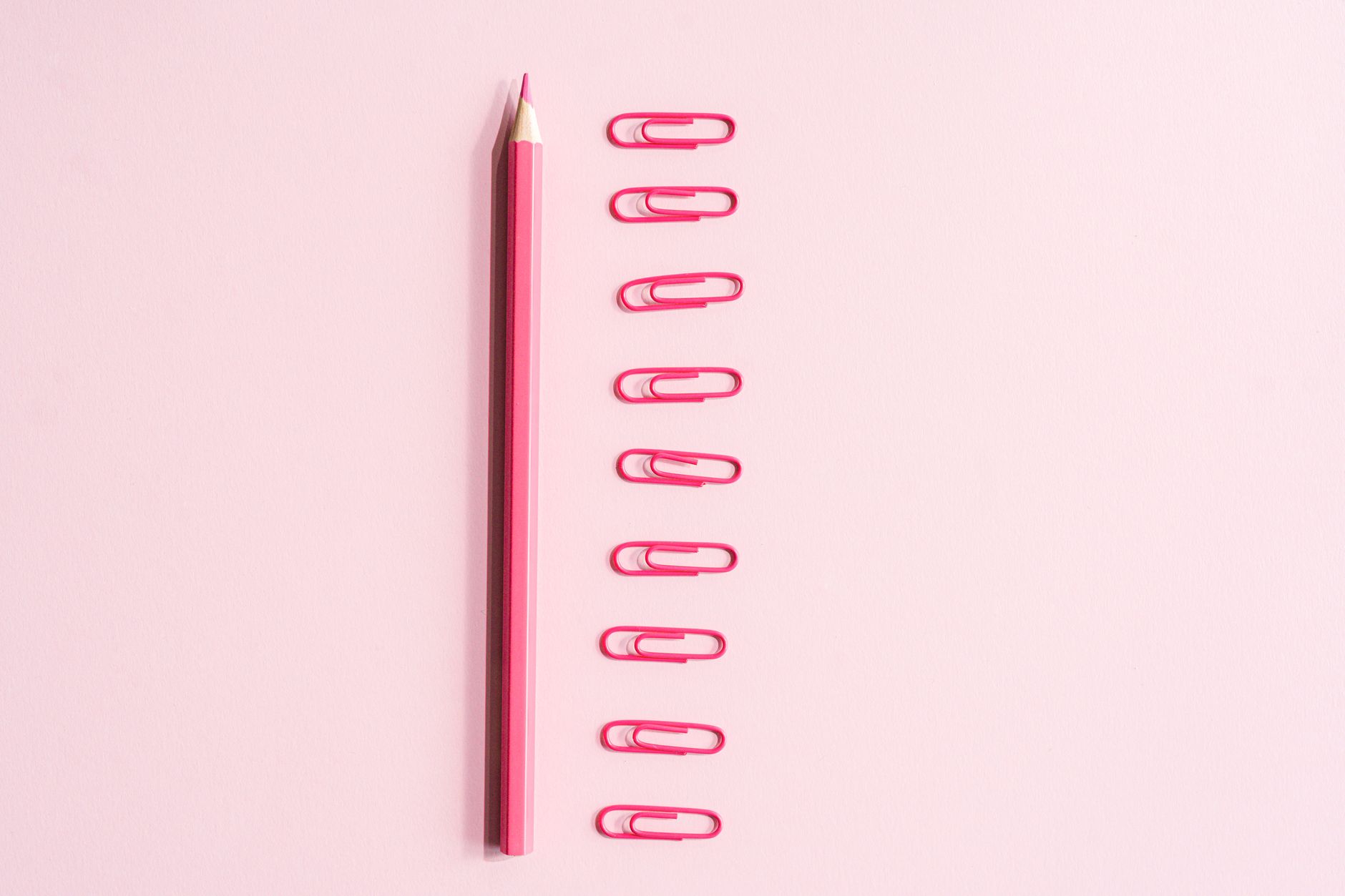 A top view of a pink pencil and pink paper clips arranged neatly on a pastel background.