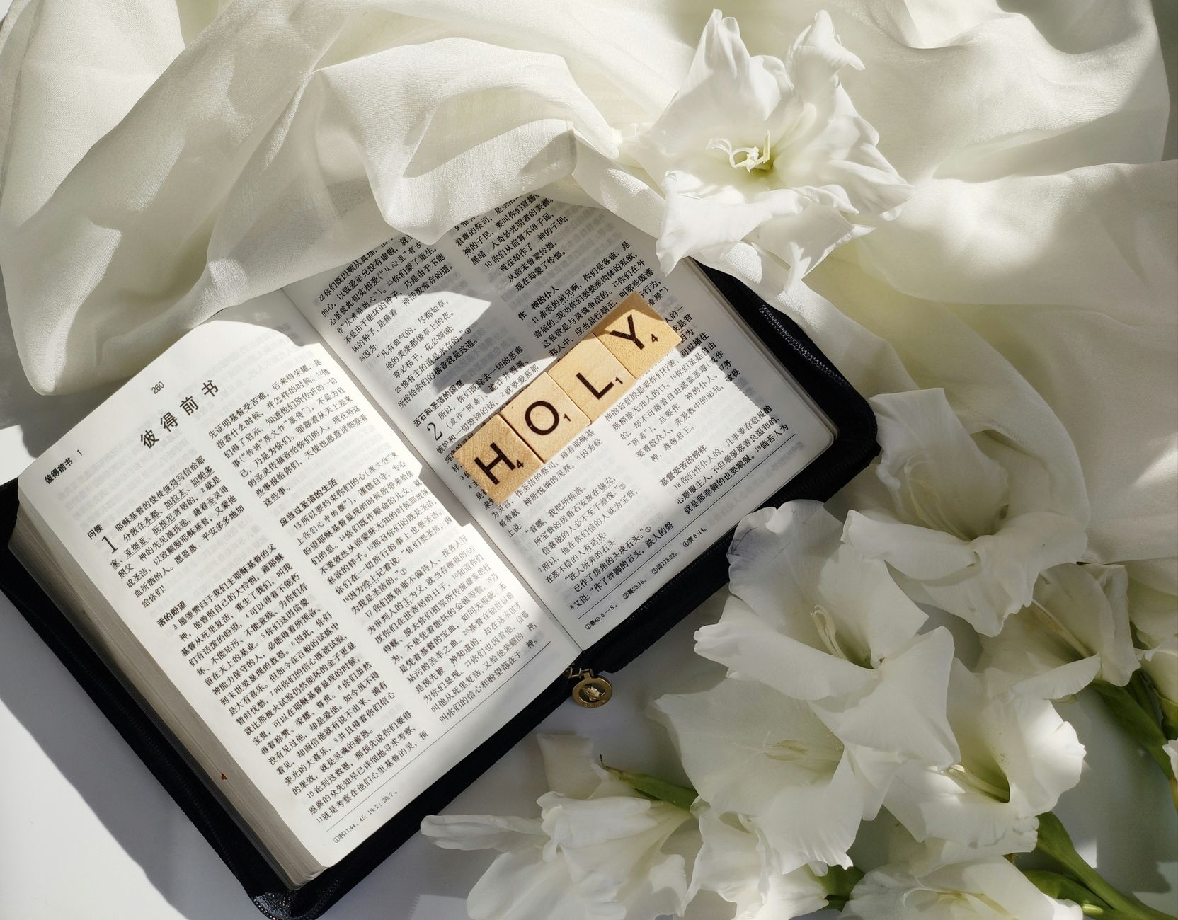 A Chinese Bible open with 'Holy' spelled in Scrabble tiles, surrounded by white flowers on a soft fabric background.