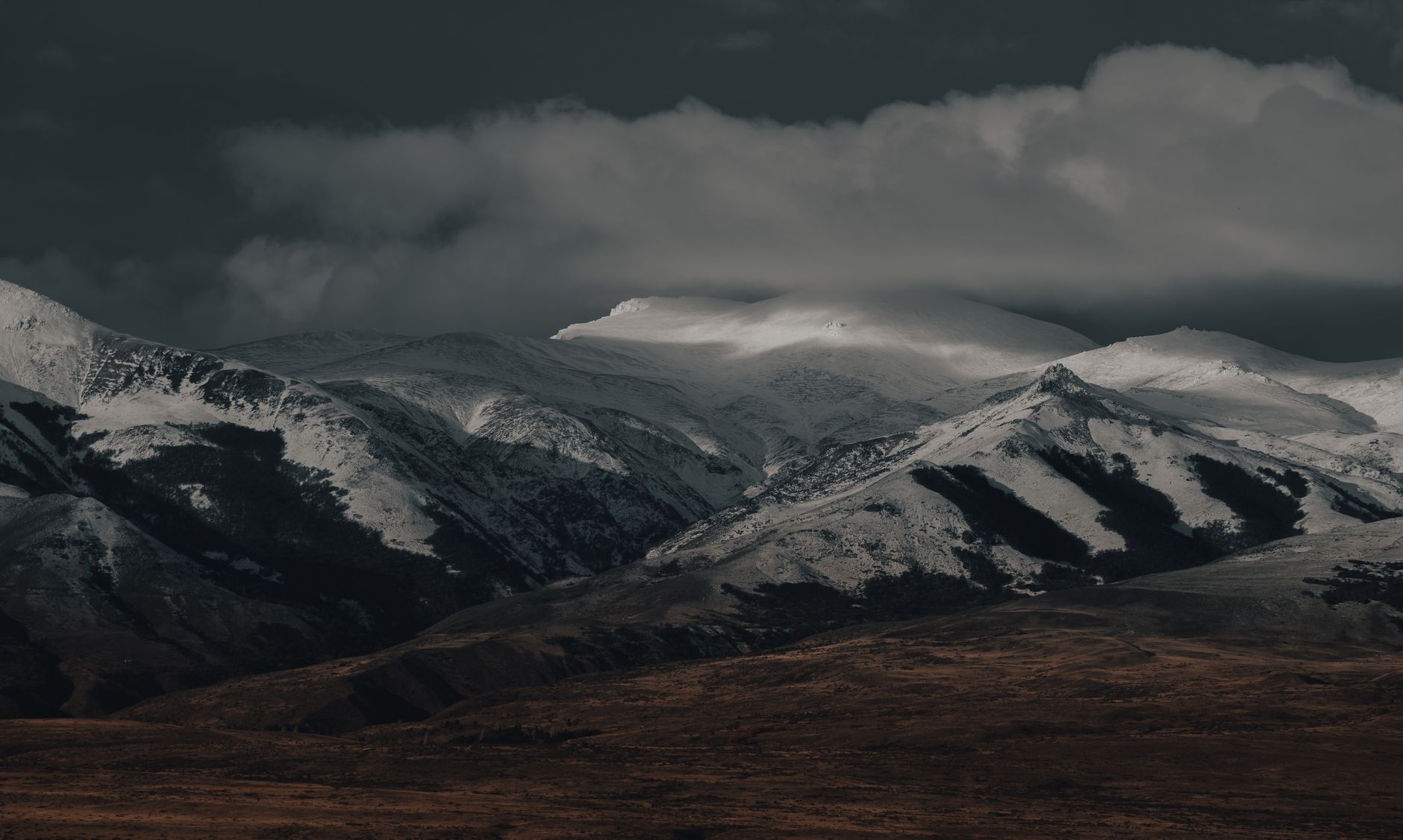 A dramatic snow-covered mountain range beneath a dark, cloudy sky, showcasing rugged natural beauty.