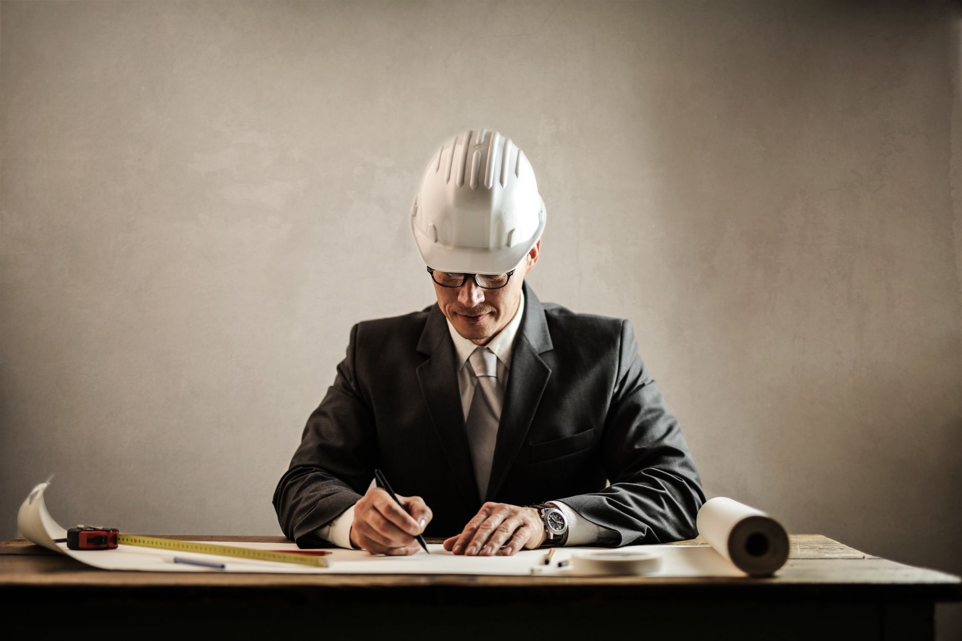 Focused young male architect in formal suit and hardhat sitting at table and drawing draft while working on project in office