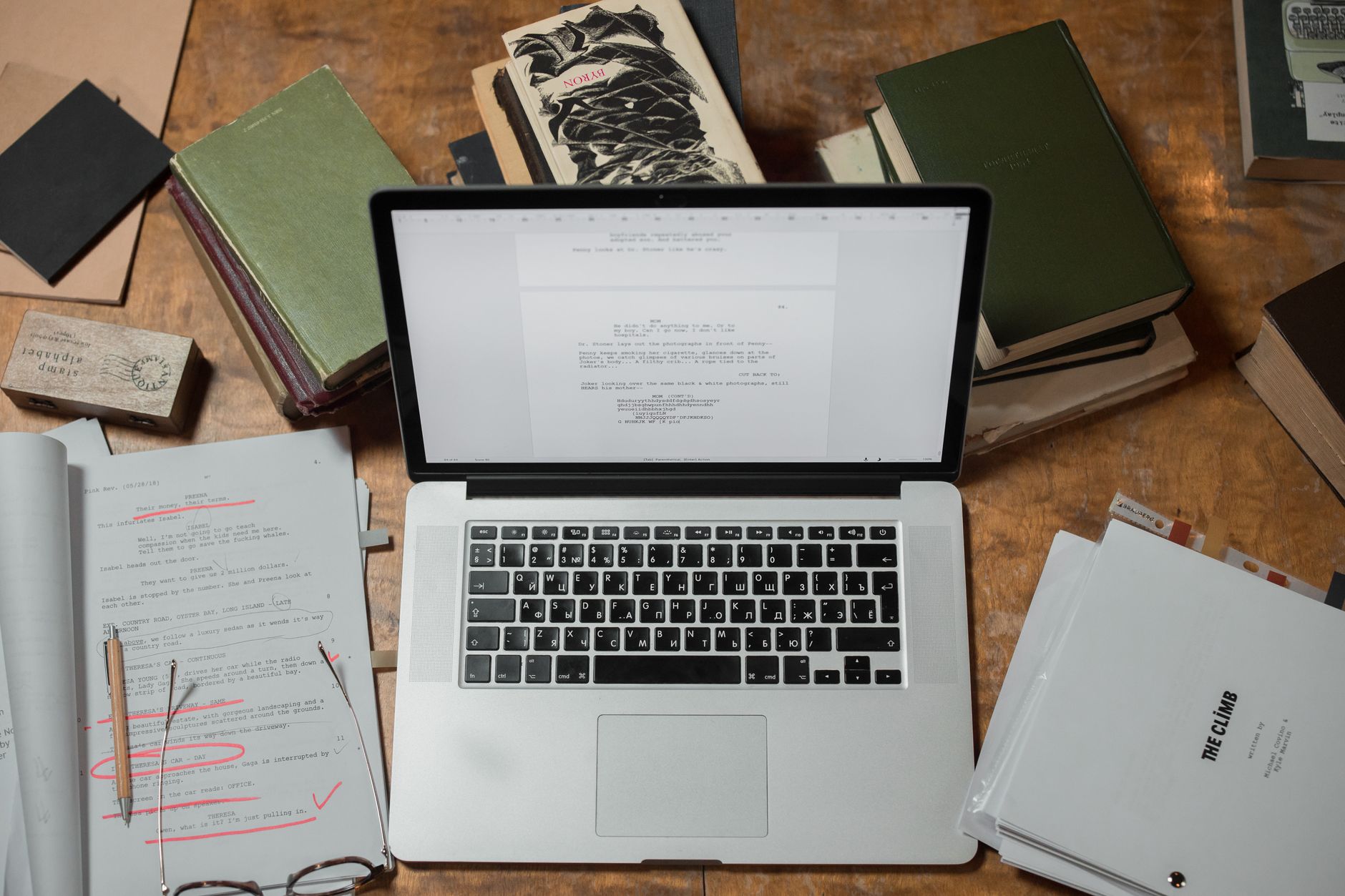 A laptop surrounded by books and scripts on a wooden desk, perfect for literature and technology themes.