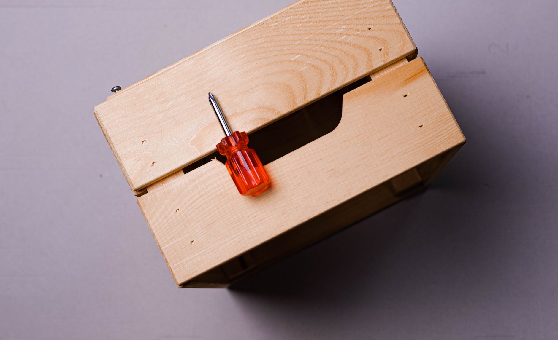 Minimalist close-up of a wooden box with a red screwdriver on a gray background.