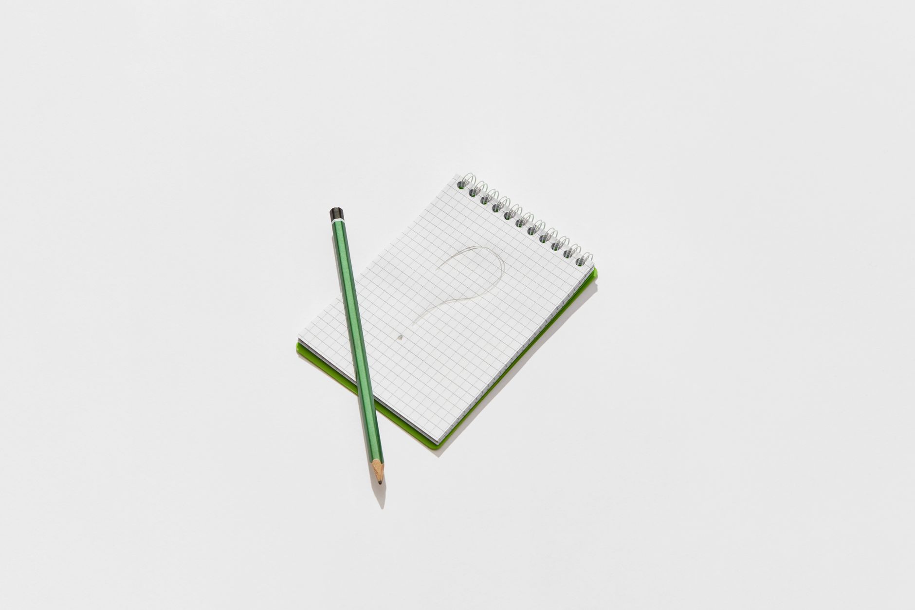 Minimalist photo of a green notepad and pencil on a white background with a drawn question mark.