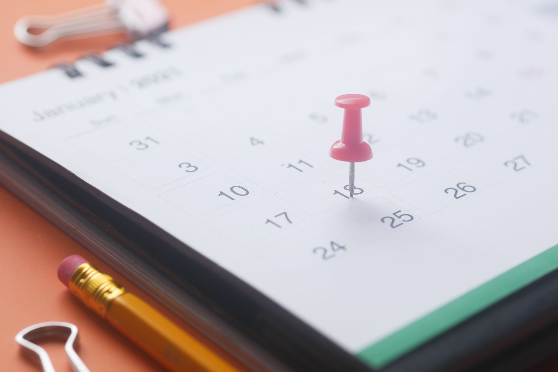 A close-up of a calendar page with a pink push pin, pencil, and binder clip on an orange background.