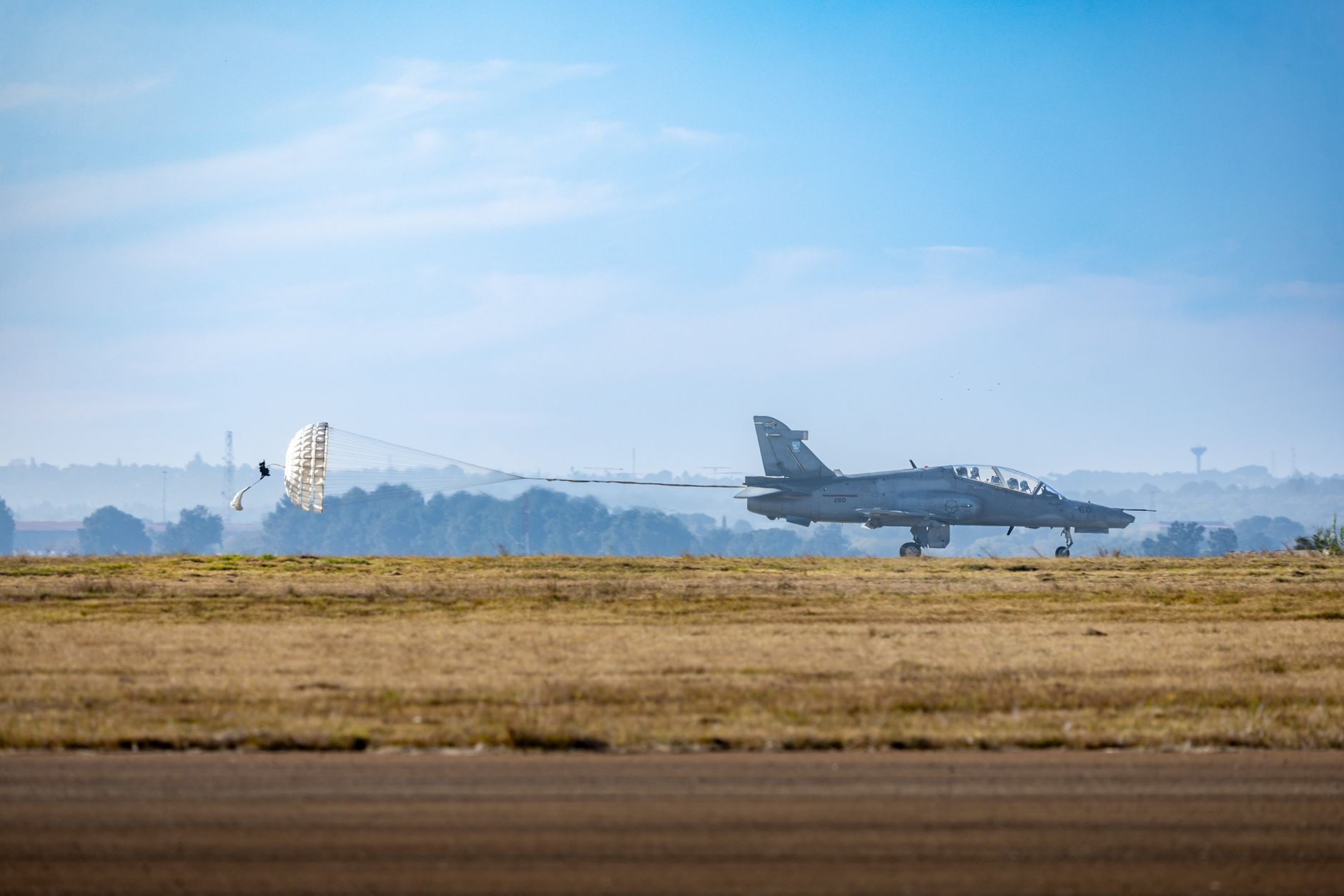 A military jet lands with a parachute deployed on a clear day at the airstrip.