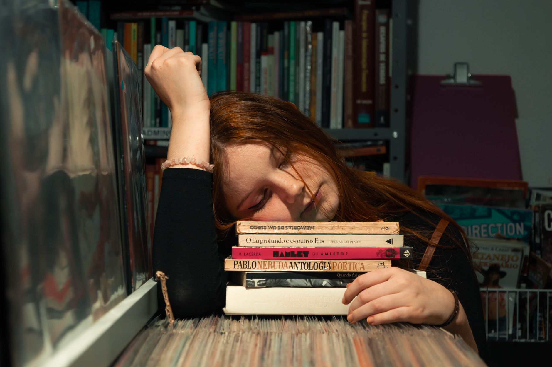 A young woman rests her head on a stack of books in a cozy library setting.