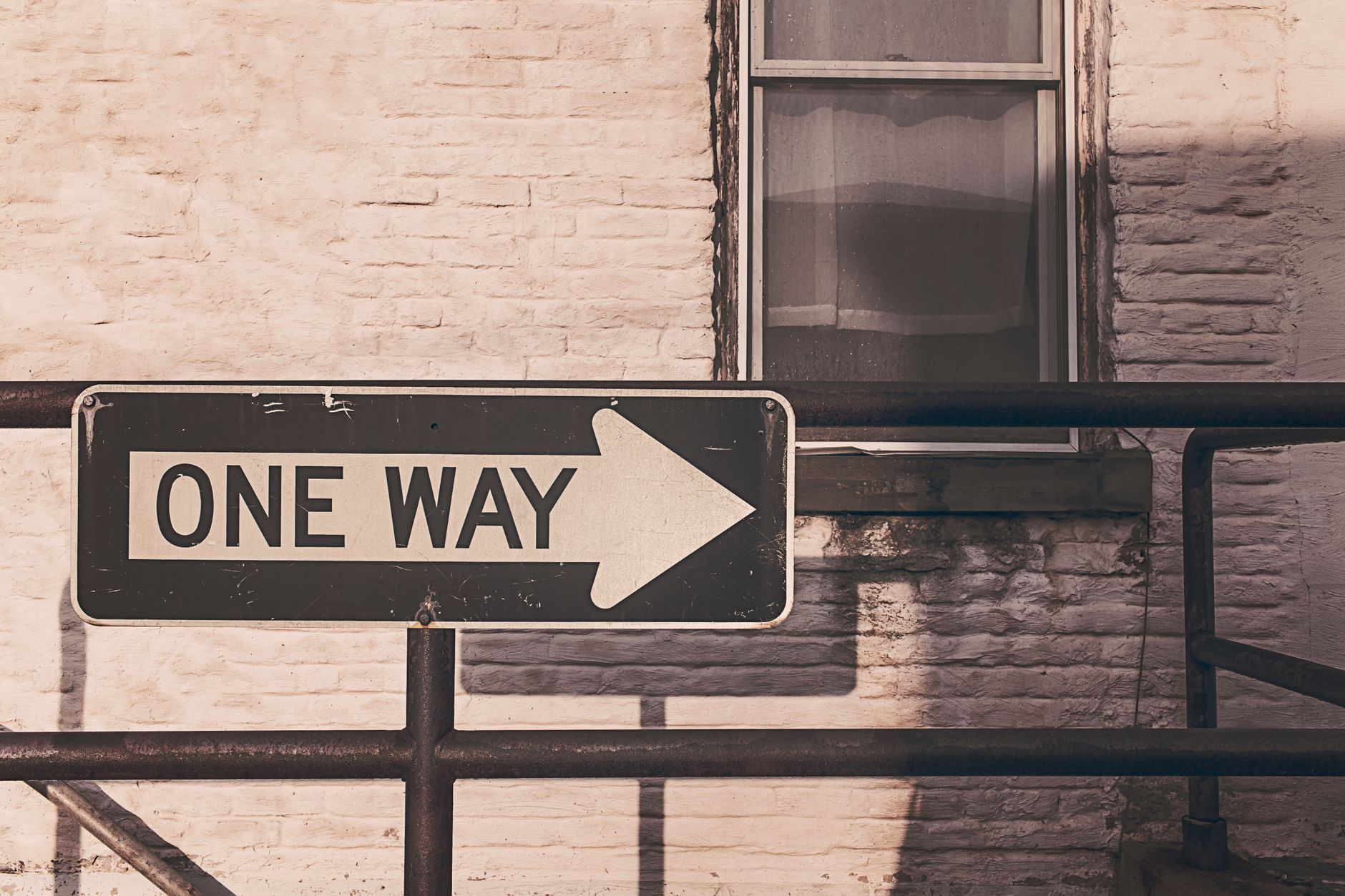 A vintage one way sign pointing right, mounted on a metal railing against a brick wall in urban setting.