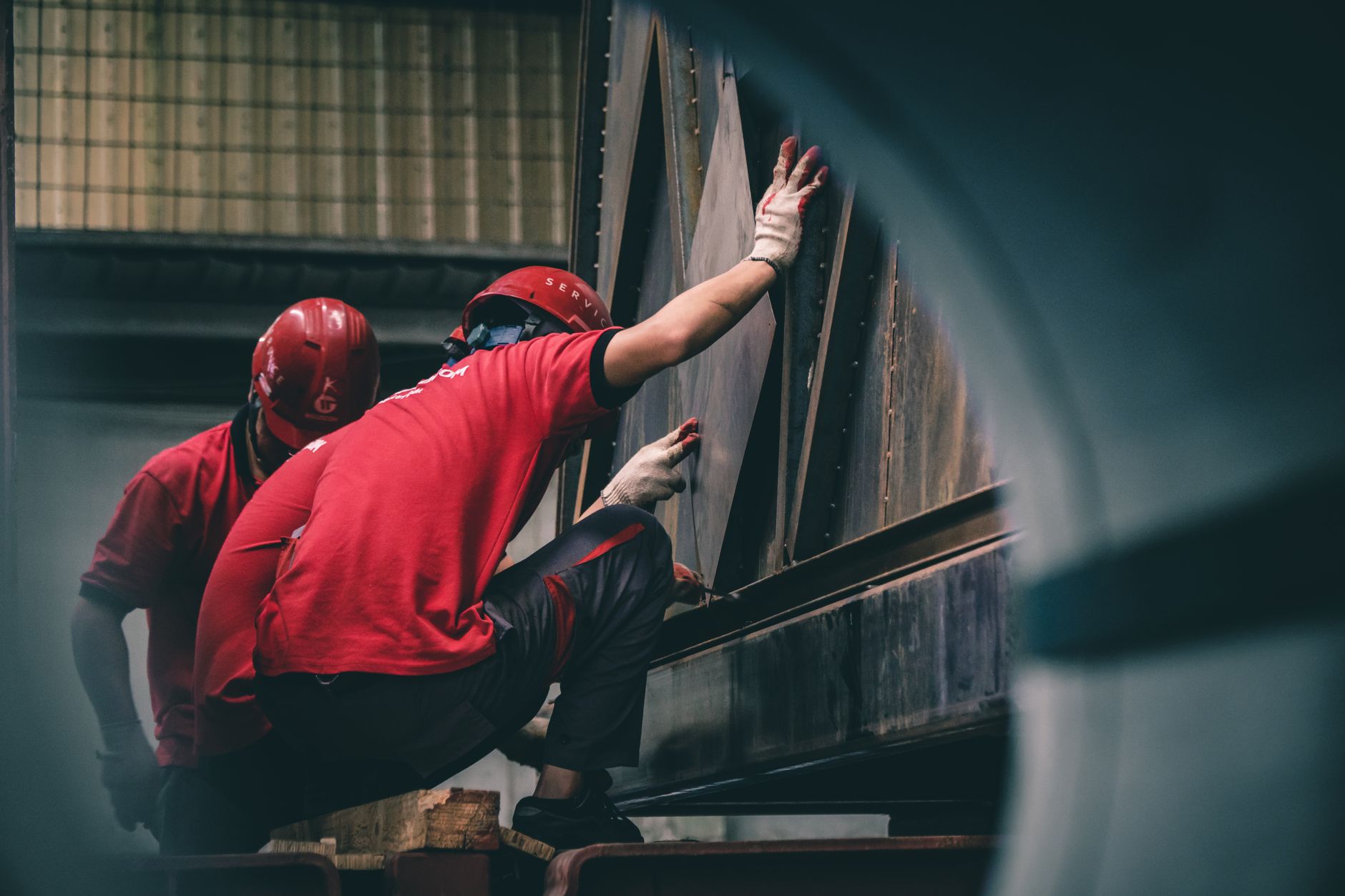 Two workers in red helmets assembling metal components inside a factory.