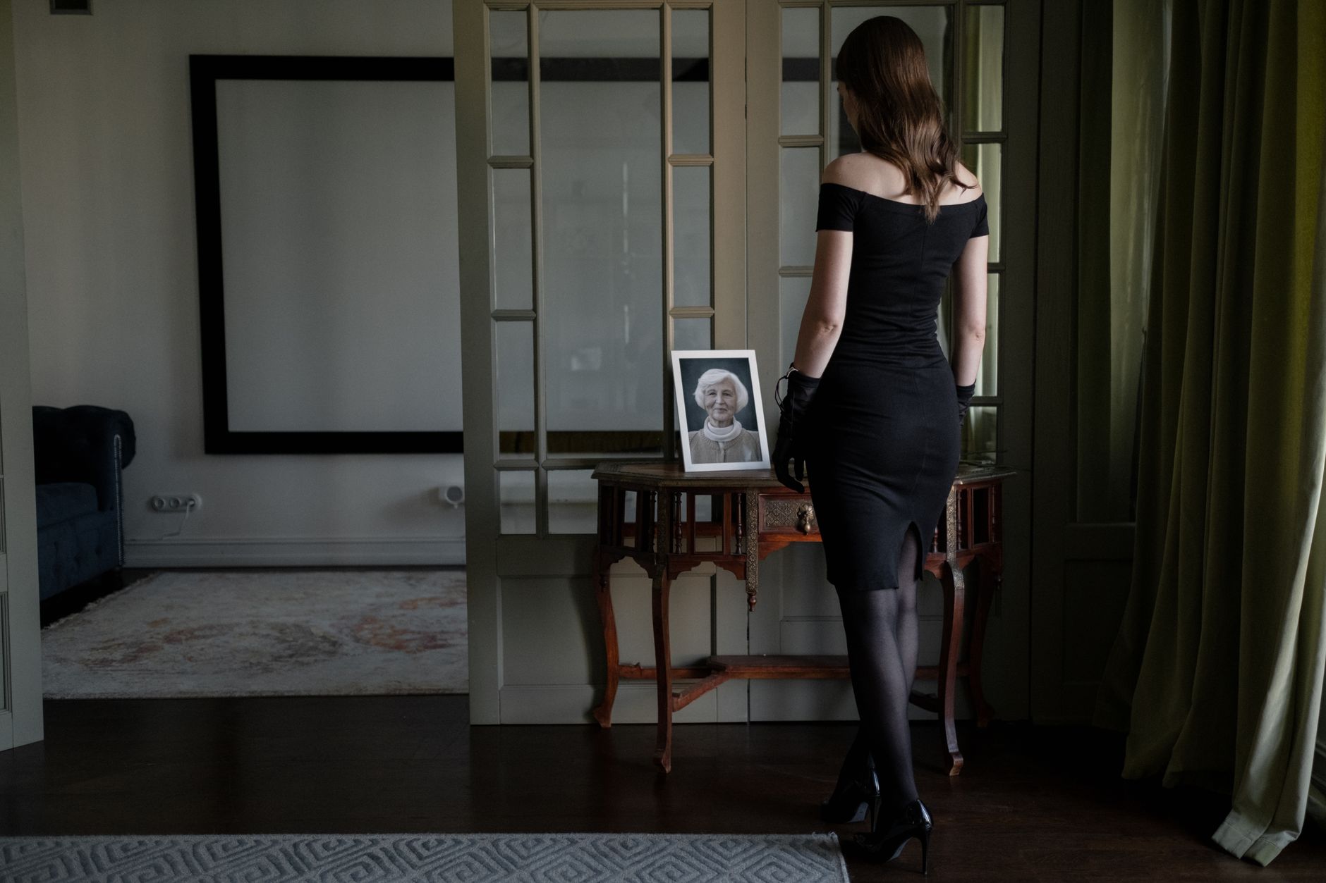 A woman in a black dress stands solemnly by a portrait, conveying a sense of loss and mourning.