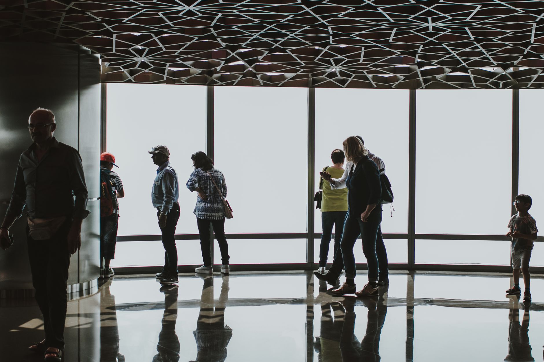 Silhouettes of tourists observing the Dubai skyline from an indoor observation deck.