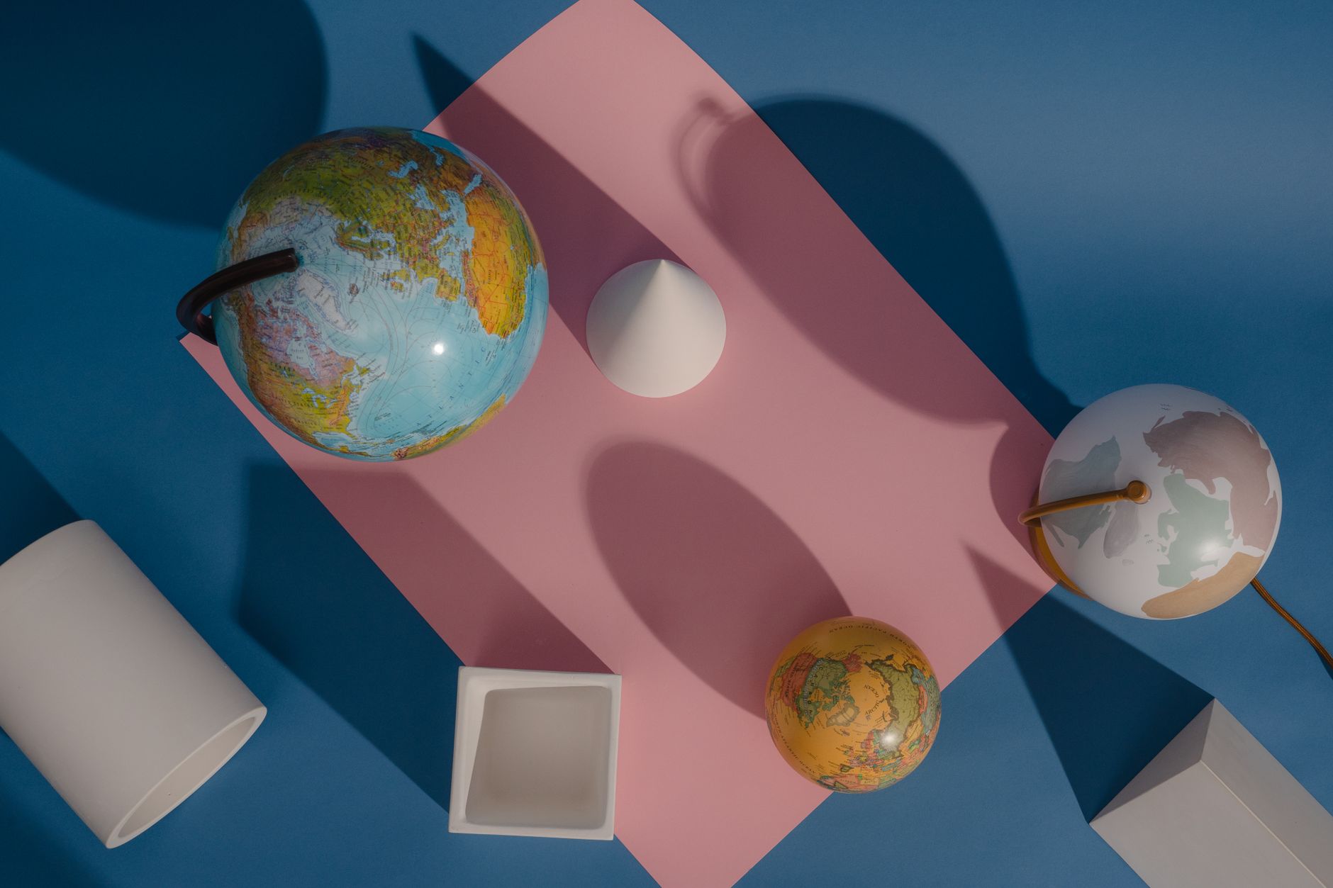 Artistic arrangement of globes and geometric shapes casting shadows on pink and blue.