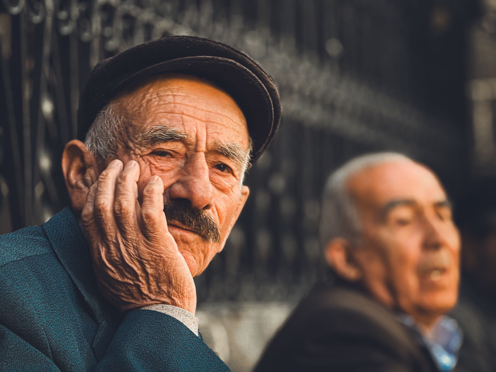 Elderly man in thoughtful pose wearing a cap, sitting outdoors in a sunny setting.