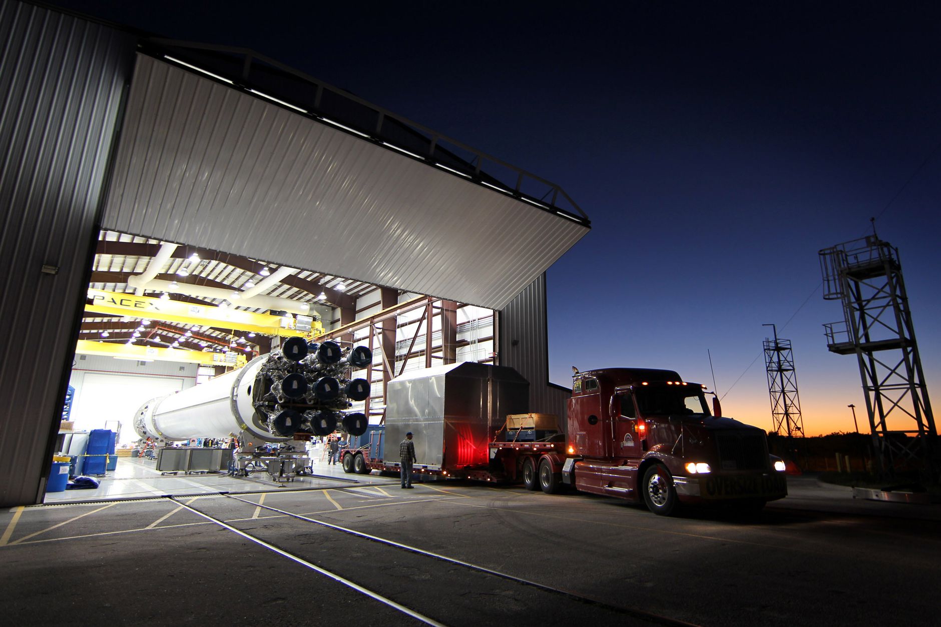 Modern cargo truck parked near hangar with massive rocket core inside at industrial factory in evening