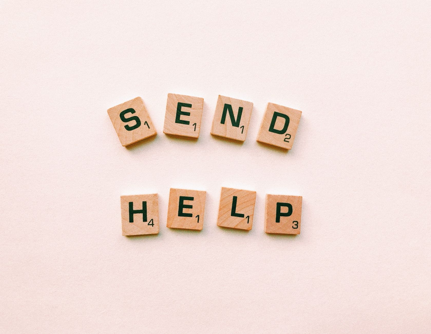 Wooden Scrabble tiles arranged to form the phrase 'send help' on a light pink background.